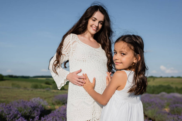 smiling girl embracing tummy of pregnant mother in openwork dress - Foto, imagen