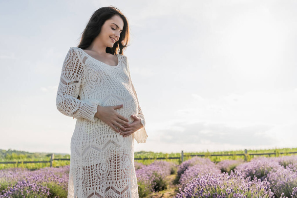 happy pregnant woman in dress touching belly in field with lavender flowers - 写真・画像