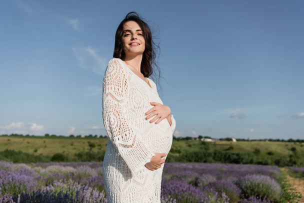 brunette pregnant woman in openwork dress smiling at camera in lavender field - 写真・画像