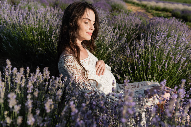 pregnant woman with long hair enjoying sitting in lavender field - Foto, imagen