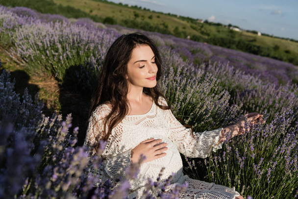 pleased pregnant woman in openwork dress sitting in lavender field - Foto, afbeelding