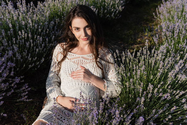 brunette pregnant woman looking at camera while sitting in meadow with flowering lavender - Fotó, kép
