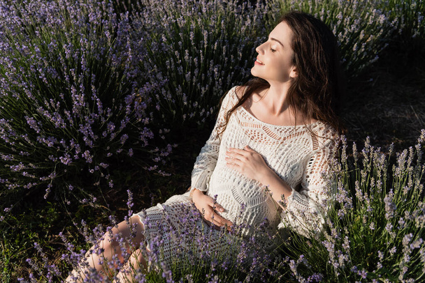 pleased pregnant woman touching tummy while sitting in lavender field - Φωτογραφία, εικόνα