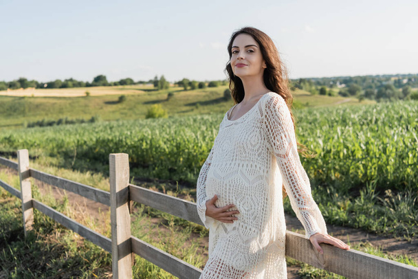 happy pregnant woman in white openwork dress standing near wooden fence in field - Photo, Image