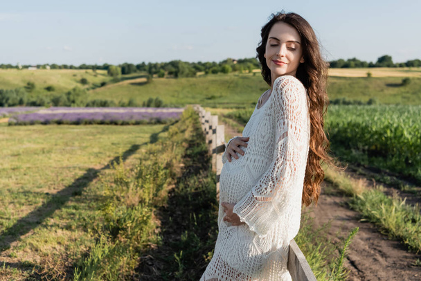 smiling pregnant woman with hands on tummy standing in summer field - Photo, Image
