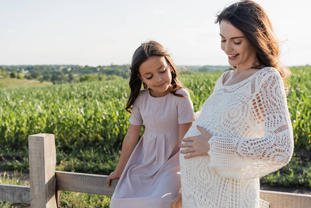 girl in pink dress sitting on wooden fence near happy and pregnant mother - Photo, Image
