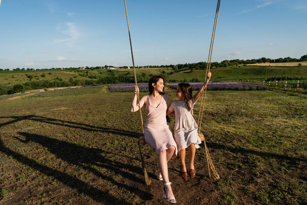 full length of woman and kid in summer dresses riding swing in meadow - Foto, Imagem