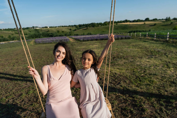 cheerful mom and daughter looking at camera while riding swing in meadow - Foto, afbeelding