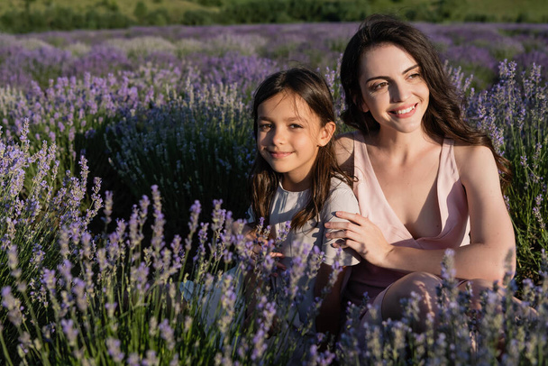 joyful brunette mother and daughter sitting in meadow with flowering lavender - Foto, Imagen