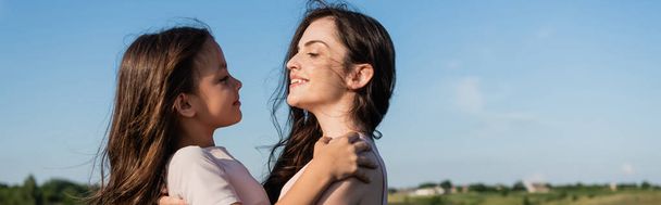 side view of happy brunette mother and daughter looking at each other outdoors, banner - Zdjęcie, obraz