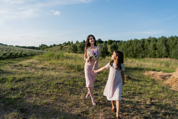 woman with bouquet holding hands with daughter while walking in field - Valokuva, kuva