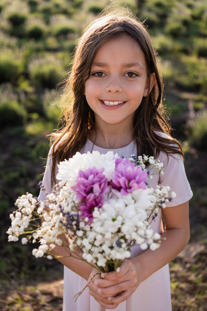 cheerful girl with floral bouquet smiling at camera in blurred field - Photo, image