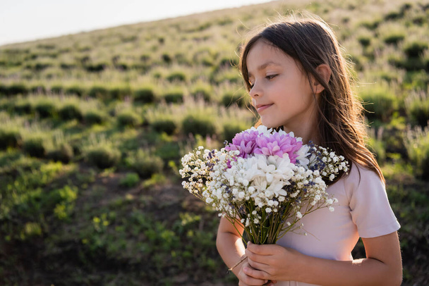 улыбающаяся женщина с длинными волосами, держащая букет цветов на летнем поле - Фото, изображение