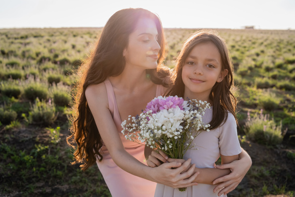 brunette woman embracing daughter holding flowers in field - Foto, Imagen