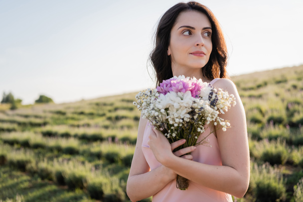 brunette woman with bouquet of fresh flowers looking away in countryside - Foto, afbeelding