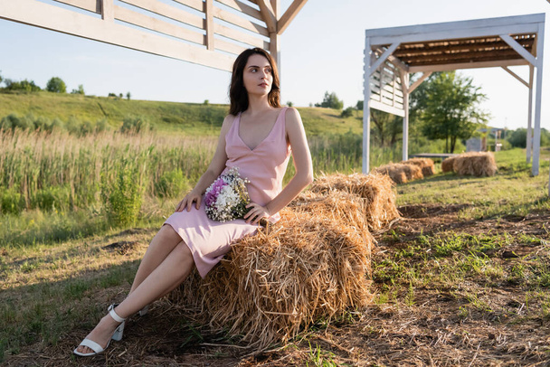 πλήρες μήκος της γυναίκας σε καλοκαιρινό φόρεμα κάθεται στα άχυρα και κοιτάζοντας μακριά - Φωτογραφία, εικόνα