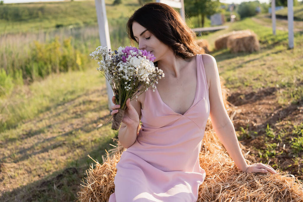 brunette woman with closed eyes sitting on haystack and smelling bouquet - Foto, Bild