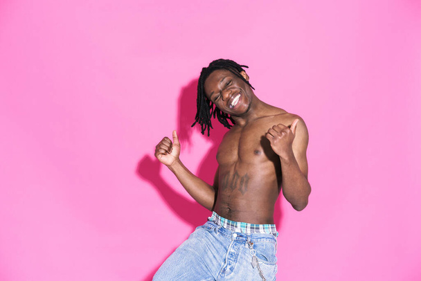Black shirtless man with dreadlocks laughing and dancing isolated over pink background - Fotoğraf, Görsel