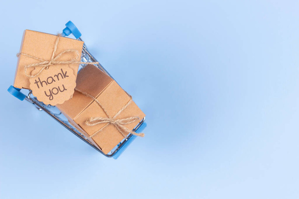 Mini shopping cart on light blue background. Kraft paper boxes with tag inscription Thank you inside trolley. Top view. E-commerce, online shopping concept. - Foto, Imagen