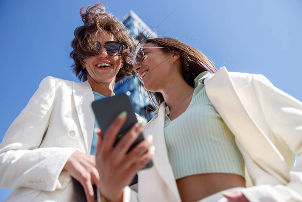 Smiling female friends in trendy outfits use phone while relaxing in city. Blurred background - Foto, afbeelding