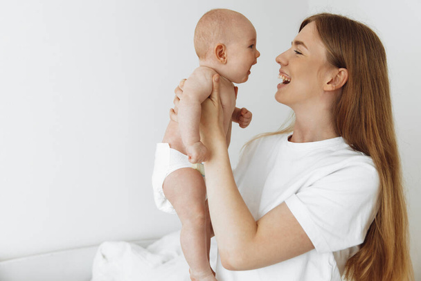 The happiness of motherhood. Portrait of a joyful mother with a cute little baby in her arms, Home portrait of a newborn with mother on the bed. Mom holds and kisses her child. loving family - Fotoğraf, Görsel