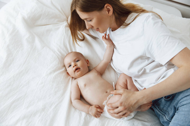 Love and tenderness, motherhood concept. Loving mother hugging her cute little baby in a diaper lying together on the bed, top view. Mom plays with the baby at home. Young women communicate with small children at home, - Фото, изображение