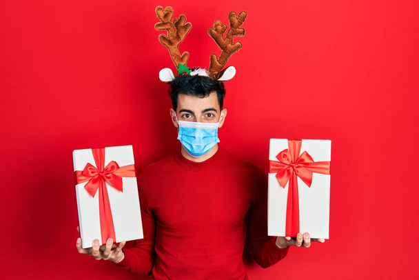 Young hispanic man wearing cute christmas reindeer horns and medical mask holding gifts relaxed with serious expression on face. simple and natural looking at the camera.  - Photo, Image