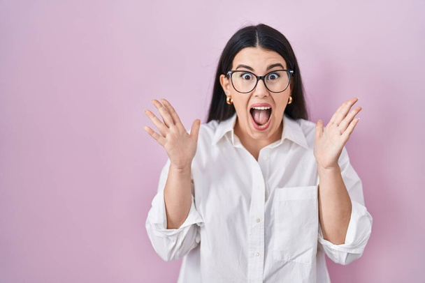 Young brunette woman standing over pink background celebrating crazy and amazed for success with arms raised and open eyes screaming excited. winner concept  - Foto, Imagen