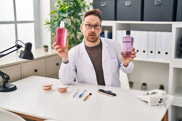 Middle age caucasian dentist man holding mouthwash for fresh breath clueless and confused expression. doubt concept.  - Foto, Bild