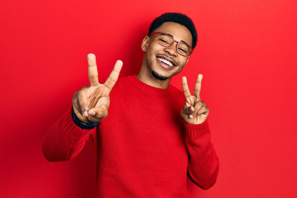 Νεαρός Αφροαμερικάνος που φοράει καθημερινά ρούχα και γυαλιά χαμογελώντας στην κάμερα που δείχνει τα δάχτυλα να κάνουν το σήμα της νίκης. Νούμερο δύο..  - Φωτογραφία, εικόνα