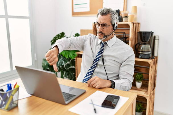 Middelbare leeftijd Spaanse zakenman die werkt op kantoor draagt operator headset kijken ongelukkig en boos tonen afwijzing en negatief met duimen naar beneden gebaar. slechte uitdrukking.  - Foto, afbeelding
