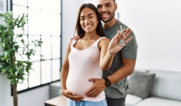 Latino-Paar umarmt sich mit Schlüssel des neuen Hauses und erwartet Baby zu Hause - Foto, Bild