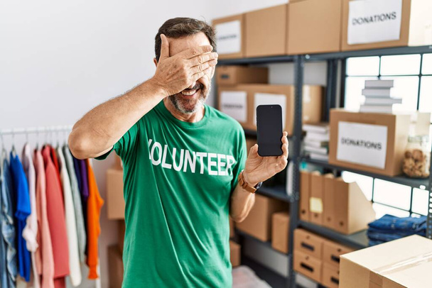Middle age man with beard wearing volunteer t shirt holding smartphone smiling and laughing with hand on face covering eyes for surprise. blind concept.  - Foto, Imagen