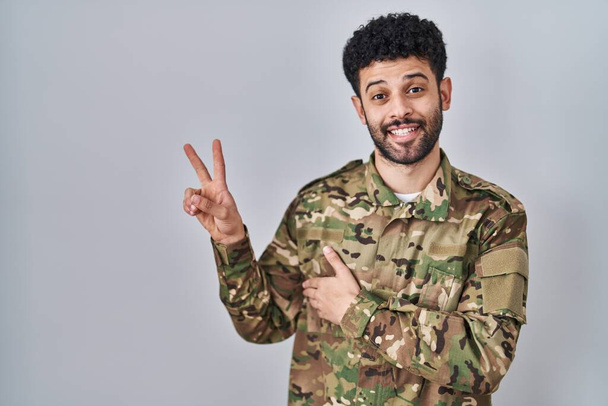 Άραβας φορώντας στολή καμουφλάζ στρατού χαμογελώντας με χαρούμενο πρόσωπο να κλείνει το μάτι στην κάμερα κάνοντας σήμα νίκης με τα δάχτυλα. Νούμερο δύο..  - Φωτογραφία, εικόνα