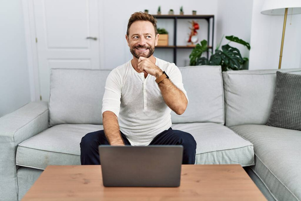 Middle age man using laptop at home with hand on chin thinking about question, pensive expression. smiling and thoughtful face. doubt concept.  - Fotoğraf, Görsel