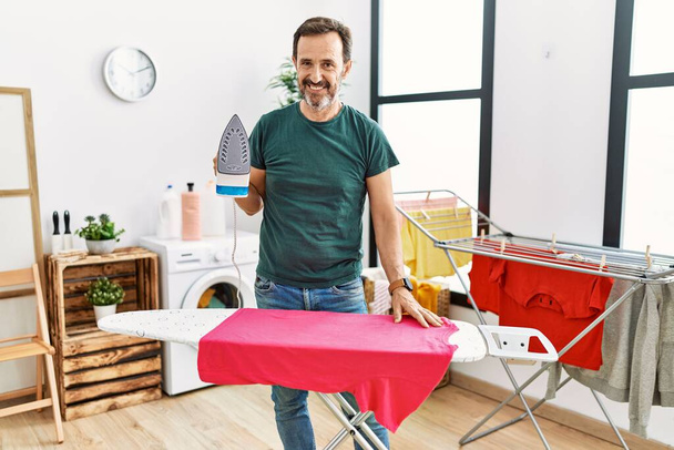 Middle age man with beard ironing clothes at home looking positive and happy standing and smiling with a confident smile showing teeth  - Fotoğraf, Görsel