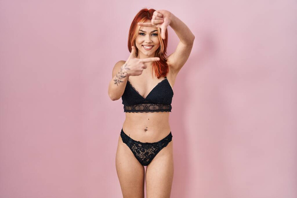 Young caucasian woman wearing lingerie over pink background smiling making frame with hands and fingers with happy face. creativity and photography concept.  - 写真・画像