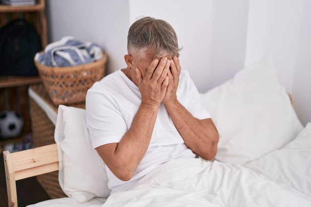 Middelbare leeftijd grijsharige man gestrest zittend op bed in de slaapkamer - Foto, afbeelding