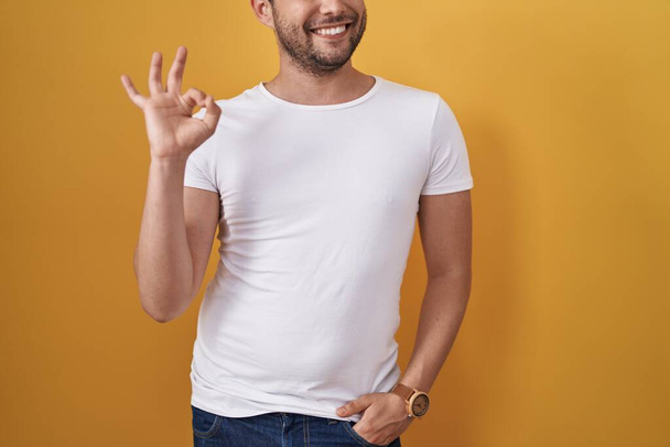 Hispanic man wearing white t shirt over yellow background doing ok sign with fingers, smiling friendly gesturing excellent symbol  - Φωτογραφία, εικόνα