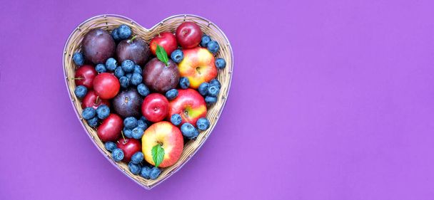Red apples, blueberry and plums in straw basket made in the shape of heart on dark shade of lavender background. Creative copy space for seasonal projects and basis designs. Close-up - Foto, immagini