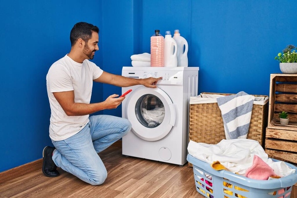 洗濯室でスマートフォンを使って洗濯機をオンに若いヒスパニック系の男 - 写真・画像