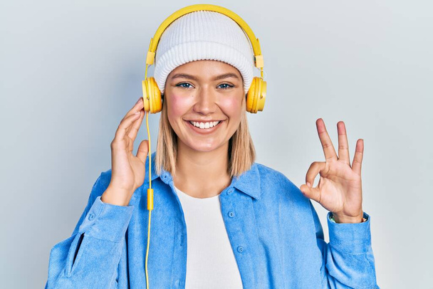 Красива блондинка слухає музику, використовуючи навушники, роблячи знак з пальцями, посміхаючись дружній жест відмінний символ
  - Фото, зображення
