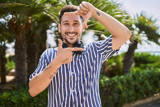 Young handsome man listening to music using headphones outdoors smiling making frame with hands and fingers with happy face. creativity and photography concept.  - Foto, Imagem