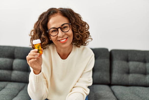 Mujer hispana de mediana edad sonriendo confiada sosteniendo pastillas en casa - Foto, imagen