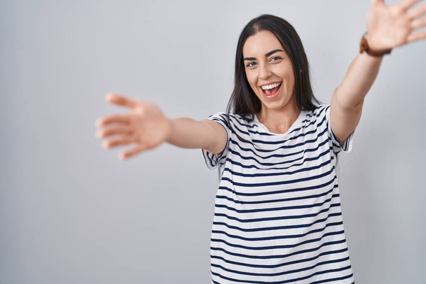 Young brunette woman wearing striped t shirt looking at the camera smiling with open arms for hug. cheerful expression embracing happiness.  - Photo, image