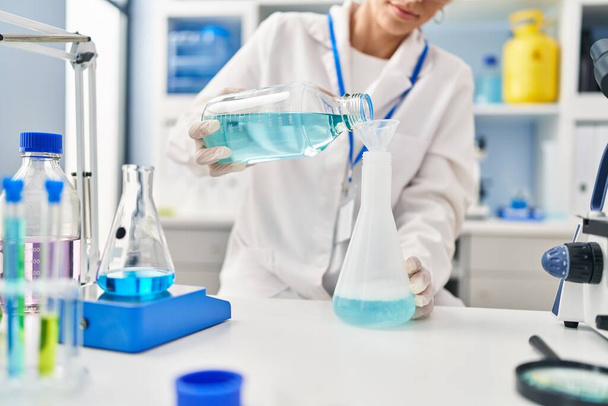 Young hispanic woman wearing scientist uniform measuring liquid at laboratory - Photo, Image