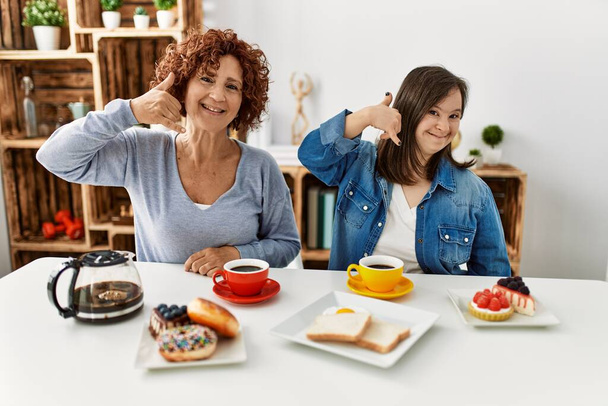 Family of mother and down syndrome daughter sitting at home eating breakfast smiling doing phone gesture with hand and fingers like talking on the telephone. communicating concepts.  - 写真・画像