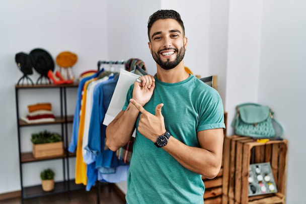 Jonge knappe man met baard die boodschappentassen vasthoudt in de winkel vrolijk met een glimlach op het gezicht wijzend met hand en vinger naar de zijkant met een vrolijke en natuurlijke uitdrukking  - Foto, afbeelding