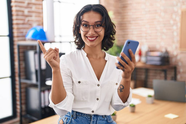 Young hispanic woman working at the office using smartphone smiling happy pointing with hand and finger to the side  - Fotoğraf, Görsel