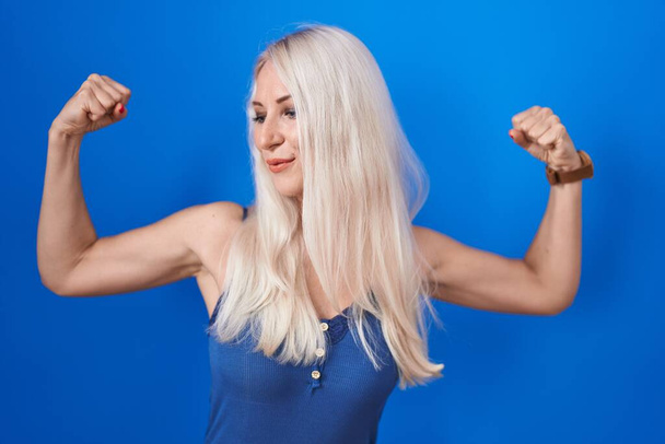 Λευκή γυναίκα στέκεται πάνω από το μπλε φόντο που δείχνει τους μυς των χεριών χαμογελώντας περήφανη. έννοια της καταλληλότητας.  - Φωτογραφία, εικόνα